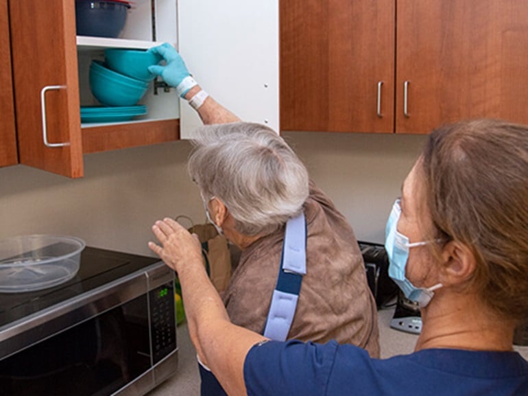Female with white hair reaching to place bowls up in a kitchen cupboard during occupational therapy session.