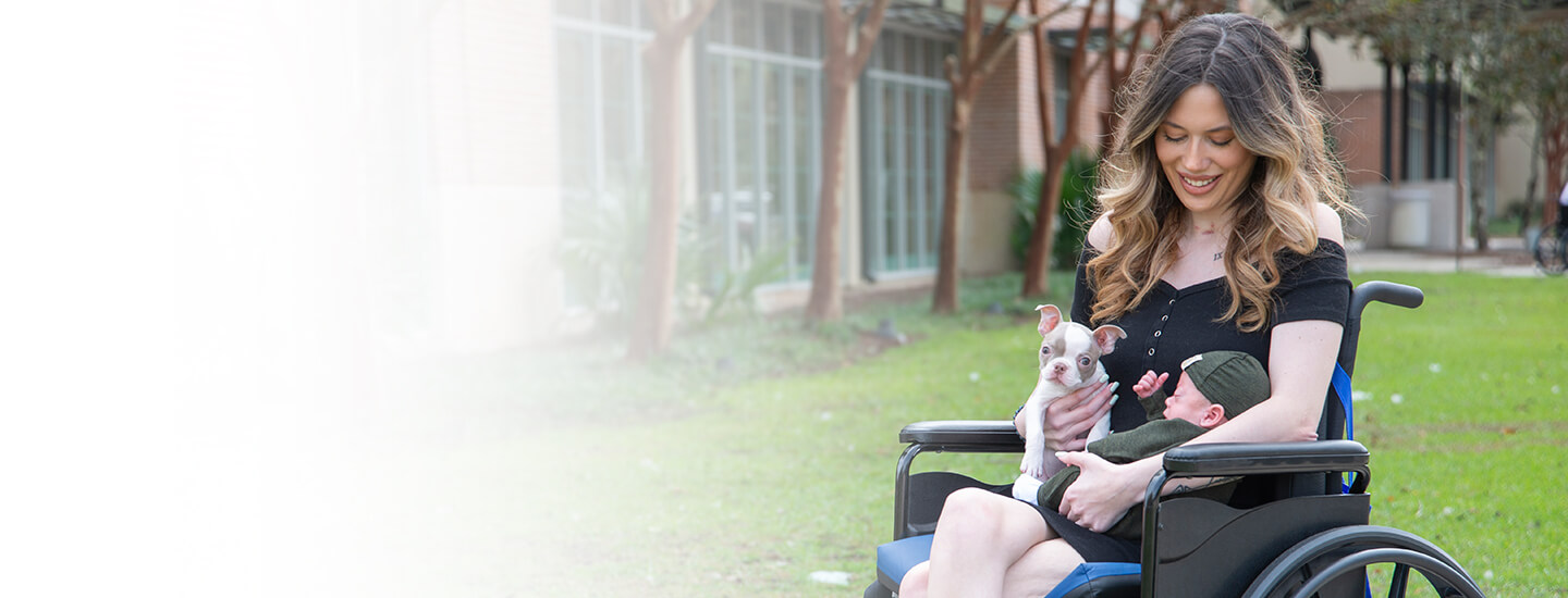 Young woman with long hair, smiling and sitting outside in a wheelchair holding a puppy and a baby.