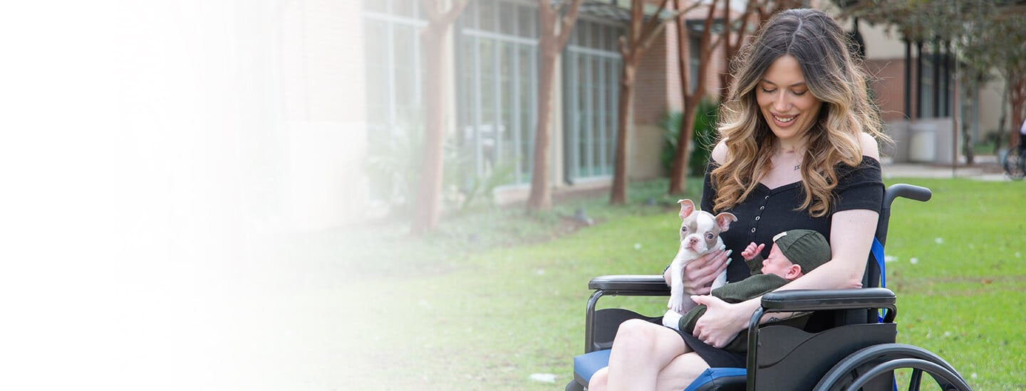 Young woman with long hair, smiling and sitting outside in a wheelchair holding a puppy and a baby.