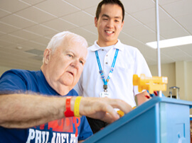 Physical therapist standing next to male patient with white hair placing small blocks in a container.