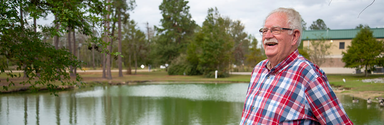 Man with white hair and wearing a red, white, and blue plaid shirt, standing in front of a small lake.
