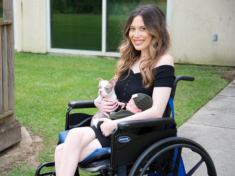 Aly seated in a wheelchair outside and holding a puppy and her son in her lap.