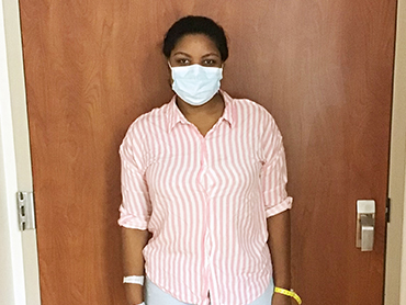 Young woman wearing a blue hospital mask and pink striped shirt standing in front of a door in a hospital.