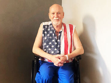 Older man smiling and wearing a Stars-and-Stripes cutoff t-shirt and sitting in a wheelchair.