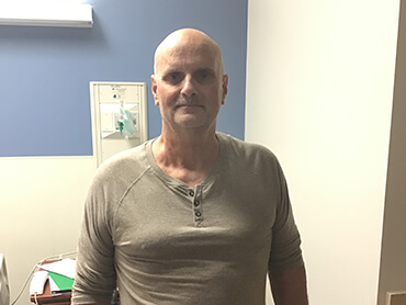 A man with a shaved head and wearing a brown pullover shirt, standing in a hospital room.