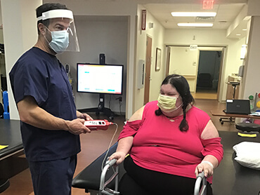 Male nurse wearing plastic face shield standing next to a female with dark braids sitting in a wheelchair in a therapy gym.