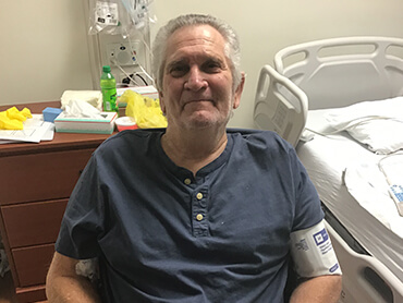 Senior male patient smiling and sitting in a chair at the side of a hospital bed.