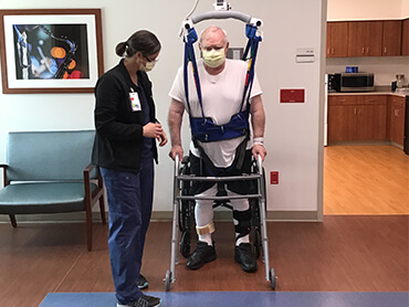 Man with white hair in a suspension safety harness and using a walker to assist in balance and walking.