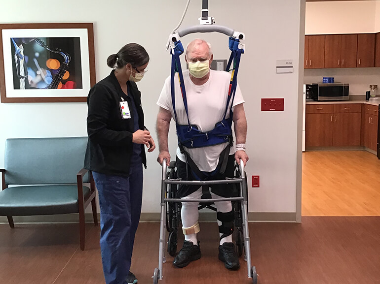Henry wearing an overhead walking harness and using a rolling walker during therapy.
