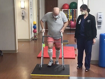 Man in a therapy gym using a walker to practice getting up and down on a step platform.