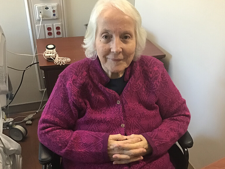 A woman with white hair and wearing a maroon cardigan, sitting in a wheelchair in her hospital room.