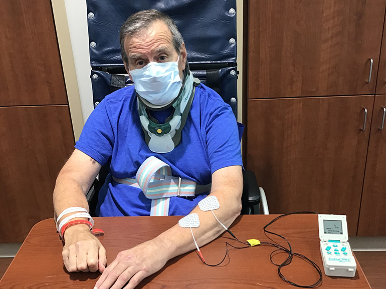 Leo wearing a cervical neck brace, sitting at a table and receiving electronic stimulation for his arm.