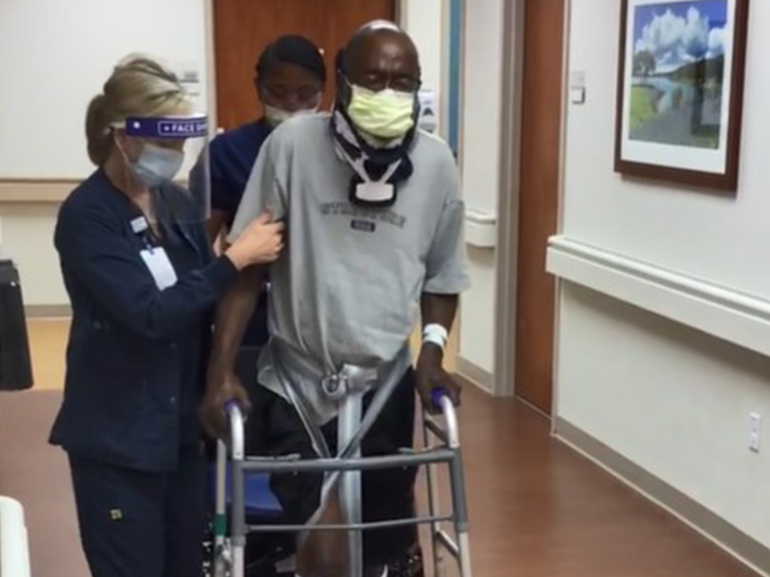 Lloyd wearing a neck brace and pushing his walker in a hospital hallway with two therapists.
