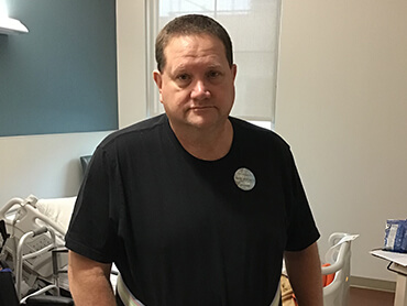 Man wearing a dark shirt standing in front of a hospital bed.
