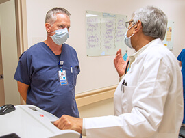 Two male doctors wearing masks and standing in a hospital hallway talking.