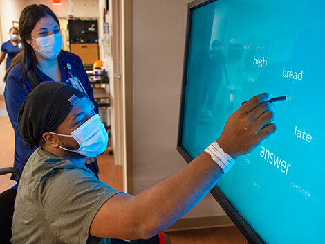 A male patient wearing a hospital mask , using a stylus to point to words appearing on a large television monitor.