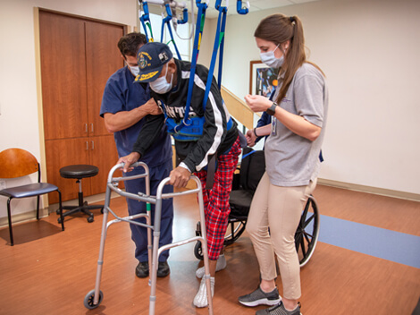 Two therapists guiding a male patient in an overhead harness and using a walker to walk.