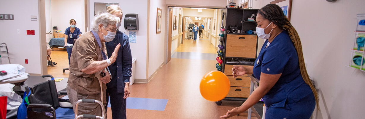 A woman with gray hair and a walker and a therapist tapping an orange balloon back and forth to each other.
