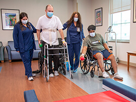 A male patient in white t-shirt and using a walker with the help of two therapists.