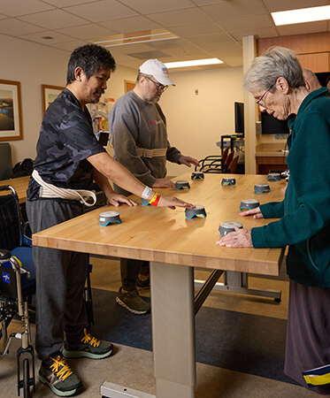 Three stroke patients standing with assistance of a gait belt at a table and reaching light up buttons