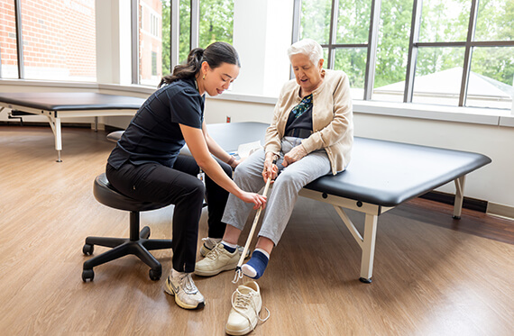 A woman is sitting on a table while a therapist shows her how to use an assistive device to put a shoe on